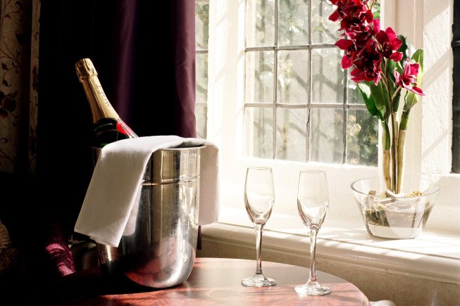 A champagne bottle in a silver ice bucket with a white napkin, two empty champagne flutes, and a vase of red flowers on a windowsill in warm