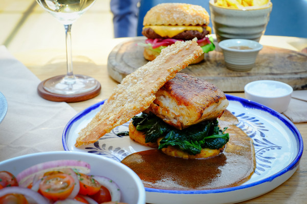 A plated fish entrée topped with a crisp garnish and sauce, served with greens and a side salad, with a burger and fries in the background.