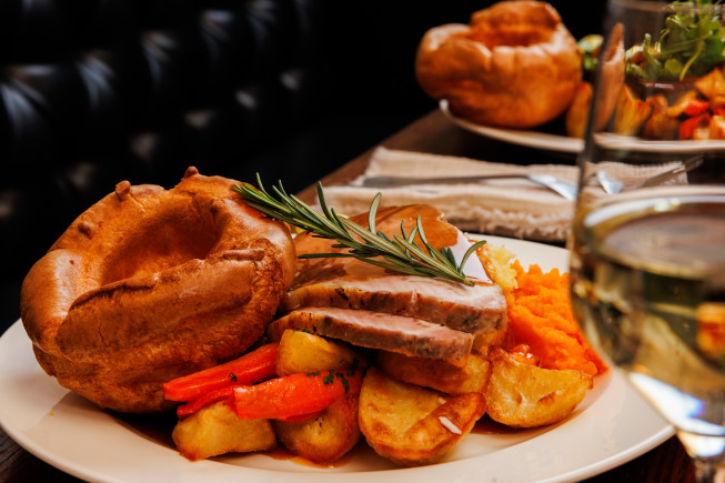 A plated roast dinner with sliced meat, Yorkshire pudding, roasted potatoes, vegetables, and a sprig of rosemary, with a glass in the right-