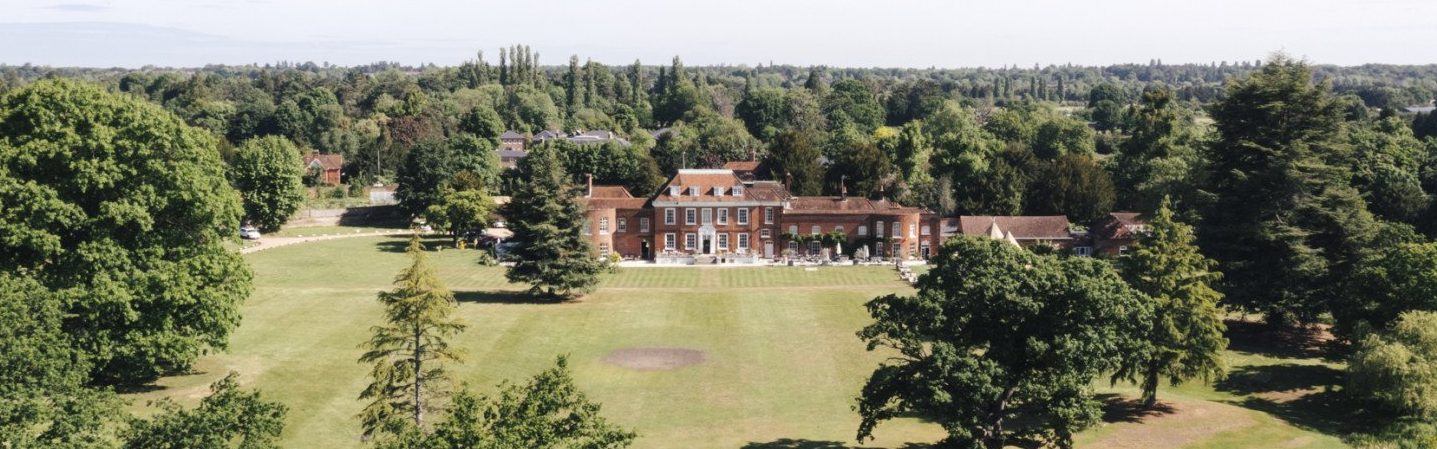 Aerial view of a large brick house set behind a broad grassy lawn, surrounded by many trees and a small body of water.