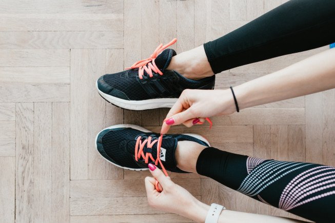 Close-up of two people’s hands tying the laces of black athletic shoes on a light wooden floor.