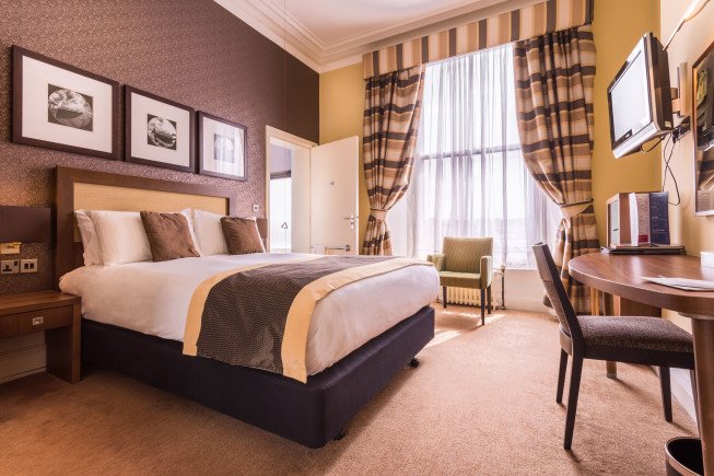 A hotel room with a large bed, desk and chair, wall-mounted TV, and a window with curtains and an armchair.