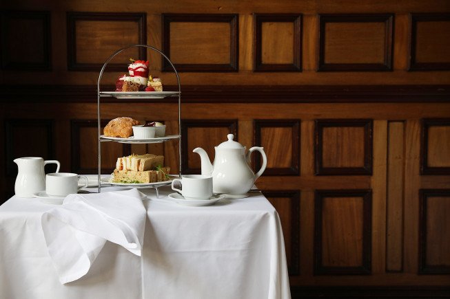 A white tablecloth-covered table with a teapot, cups, plates, and a tiered stand holding pastries and sandwiches in front of a wooden panele