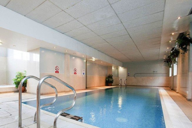 An indoor swimming pool with a metal handrail, tiled deck, and windows along one side.