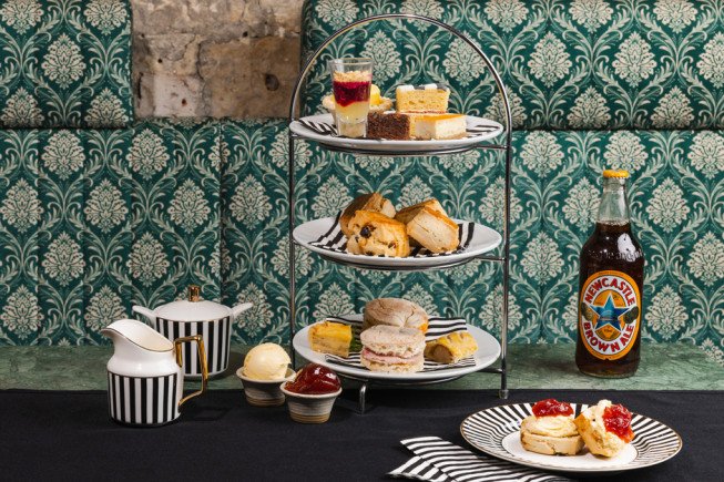 Tiered stand and plates of assorted cakes, scones, and jam alongside striped teaware and a beer bottle on a table.