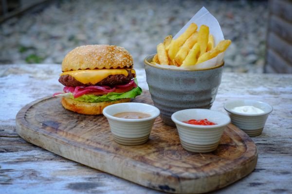 A cheeseburger with lettuce and onions sits beside a cup of French fries and three small dipping sauces on a wooden board.