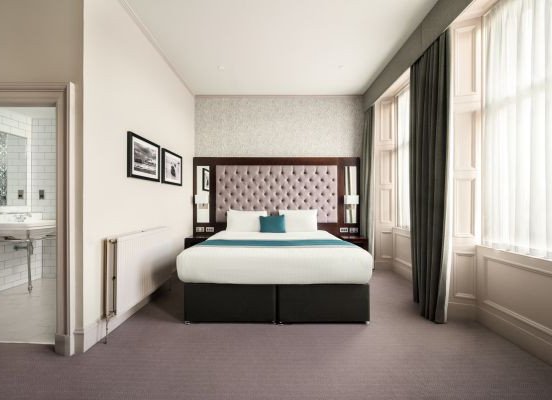 A neatly made bed with a tufted headboard in a bright hotel-style room, with large windows on the right and a bathroom visible through an aj