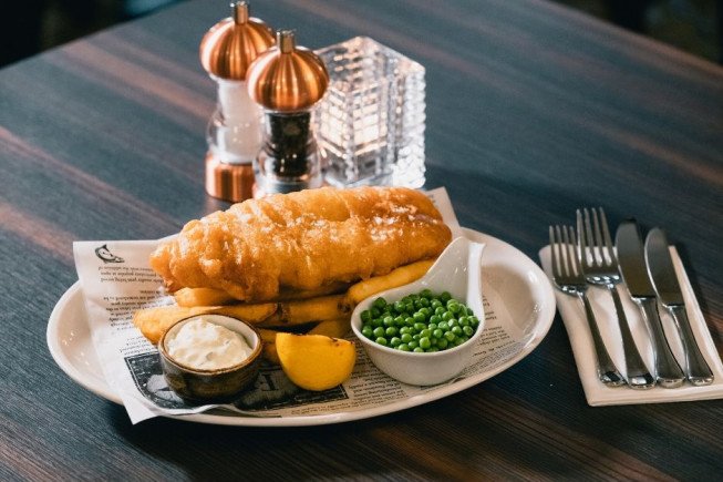 A plate of fried fish and chips with peas, tartar sauce, and lemon on a table beside cutlery and condiments.
