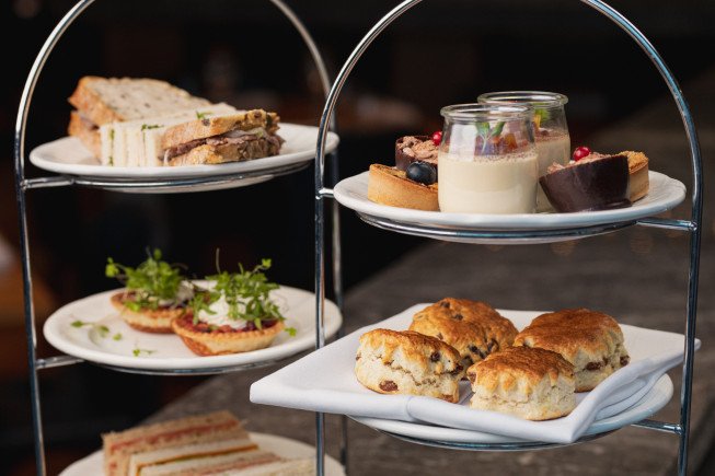 Tiered serving stand holding sandwiches, small desserts, a dessert cup, and scones on white plates.
