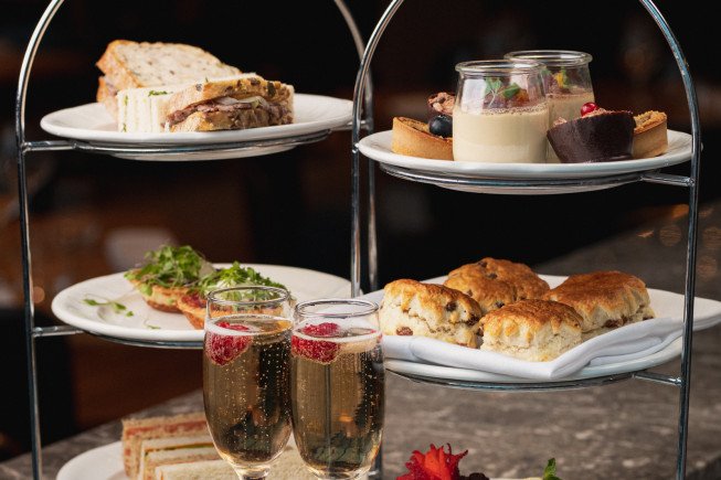 A tiered serving stand holding assorted sandwiches, pastries, cakes, and two glasses of sparkling drink.