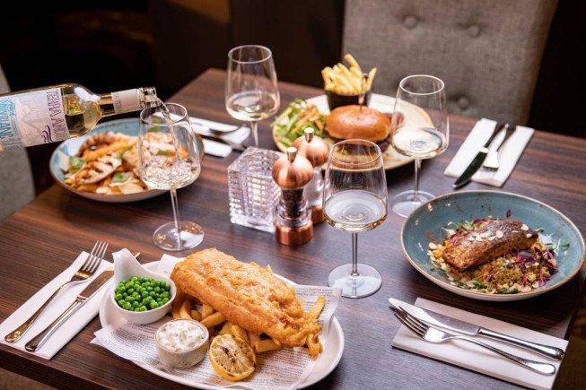A dining table set with plated fish and chips, a fish entree with vegetables, peas, condiments, wine glasses, and a bottle being poured.