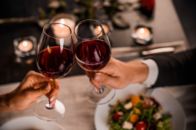 Two people clink glasses of red wine over a dining table with candles and a plate of salad nearby.