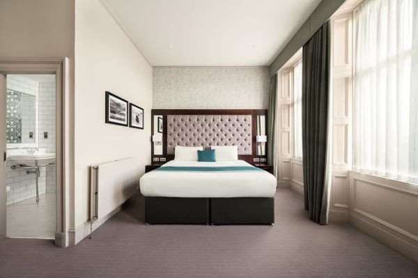 A neatly made bed centered in a modern hotel-style bedroom with a tufted headboard, framed wall art, and tall windows with curtains along a