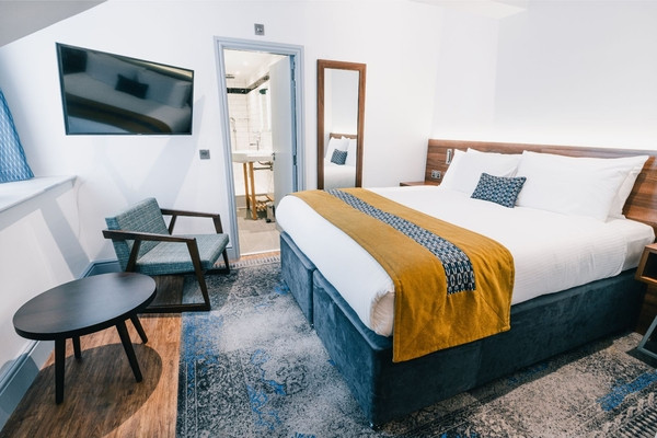 A neatly made bed with white linens and a mustard throw sits in a modern hotel room with a wall-mounted TV, chair, small table, and an open,