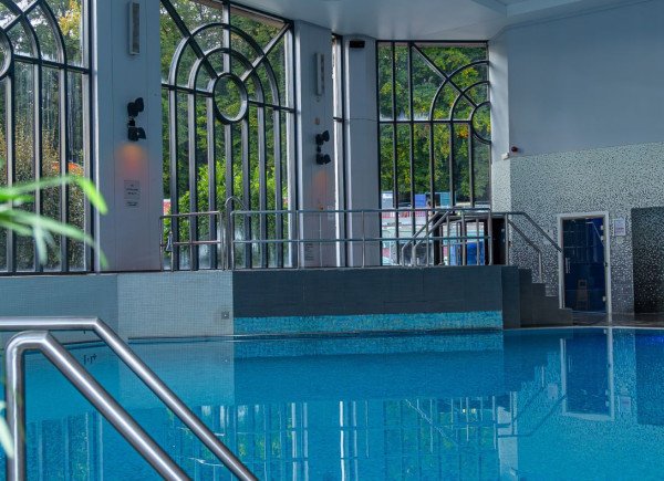 An indoor swimming pool with calm blue water, metal handrails, and large arched windows letting in daylight.