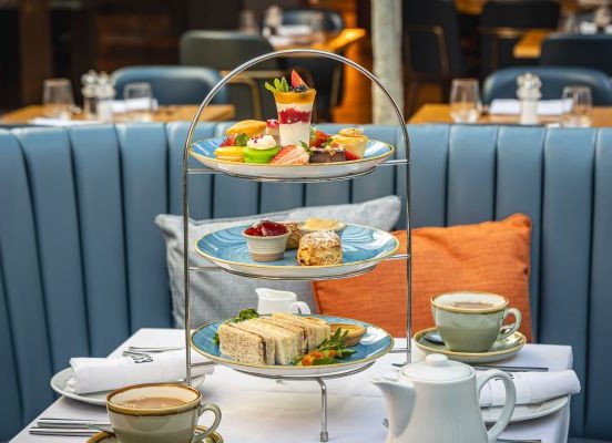 A three-tier stand with desserts and finger sandwiches on a table set with teacups and a teapot in a restaurant booth.