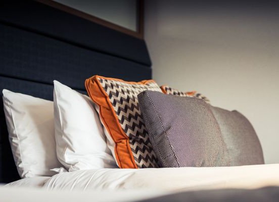 A row of white, patterned, and orange-trimmed pillows arranged against a dark headboard on a bed.