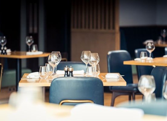 An empty restaurant interior with small wooden tables set with wine glasses, water glasses, plates, and salt and pepper shakers.