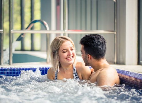 A smiling couple sits in a bubbling hot tub, facing each other and talking.