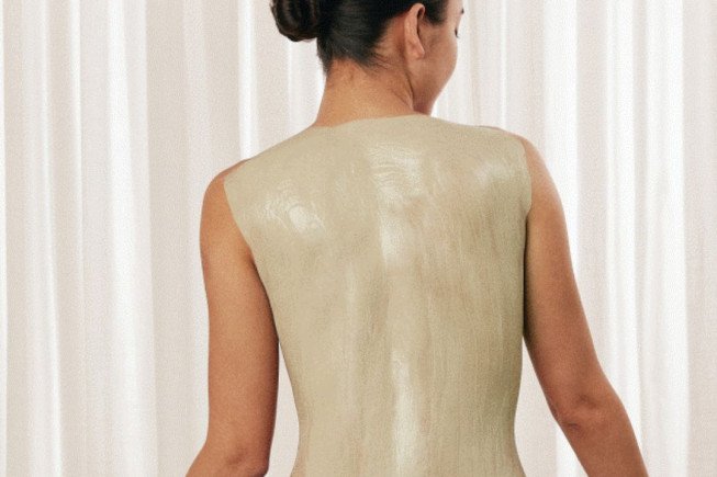 Rear view of a person wearing a sleeveless, backless light-colored dress in front of curtains.