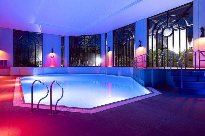 An indoor swimming pool lit with blue and pink lights, with metal handrails, steps, and large decorative windows along the walls.