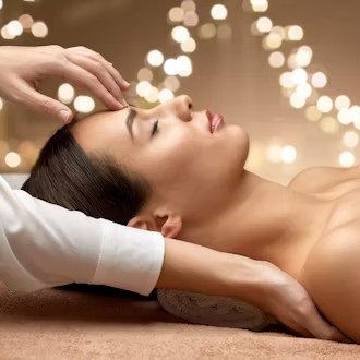 A person lying on a spa treatment table with eyes closed while a practitioner gently touches the forehead and neck, with warm string lights.