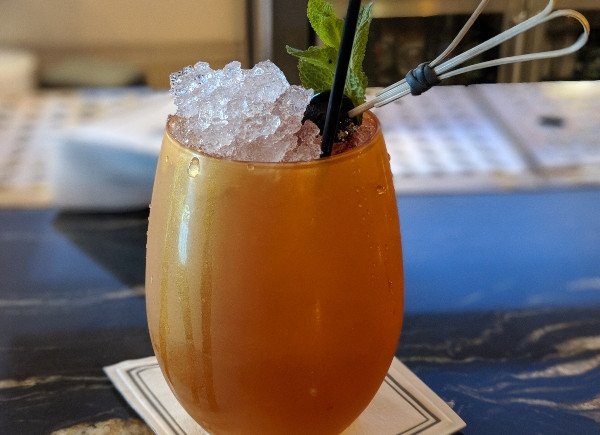 A stemless orange cocktail topped with crushed ice, mint, and a black straw on a table.
