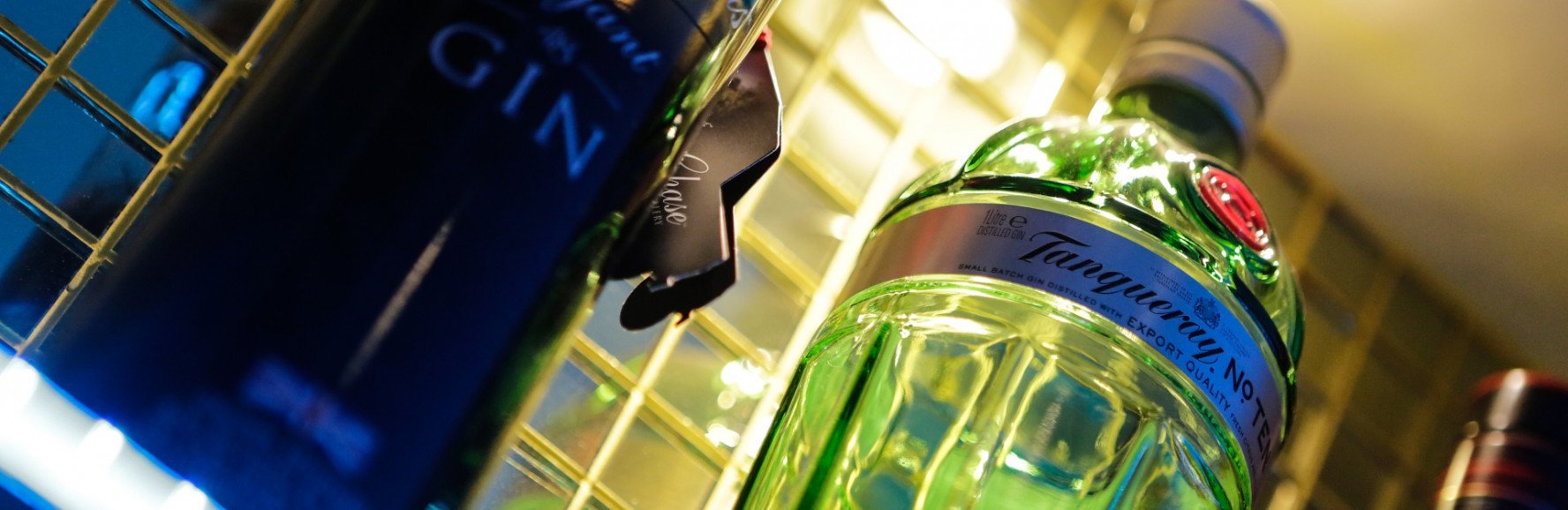 Close-up view of several liquor bottles on a shelf, including a green gin bottle and a dark blue bottle, under warm lighting.