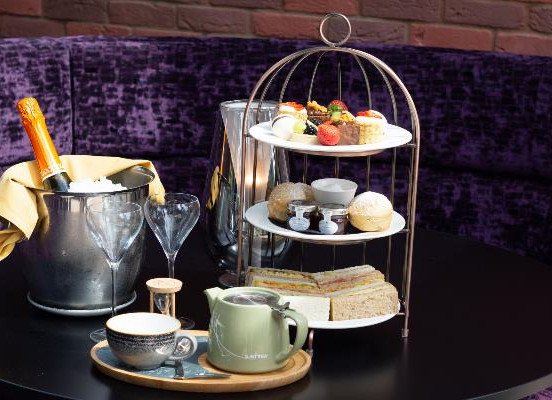 A table set with a three-tier stand of pastries and sandwiches, two empty champagne flutes, a bottle in an ice bucket, and teacups with a te