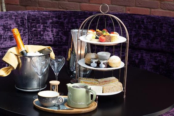 A table set with a champagne bucket, two empty wine glasses, a three-tier stand of pastries and sandwiches, and a teapot with a teacup.