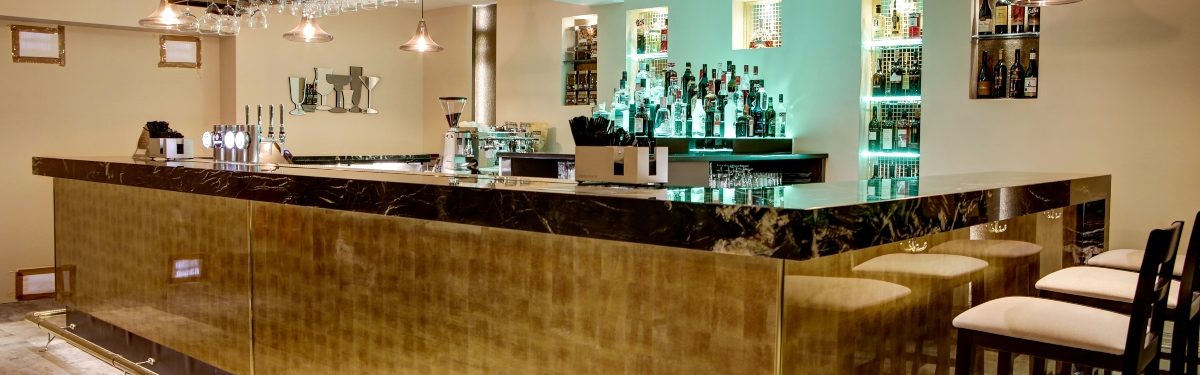 A stylish indoor bar with a gold-toned front, dark marble countertop, hanging stemware rack, pendant lights, and bottles displayed on back-s
