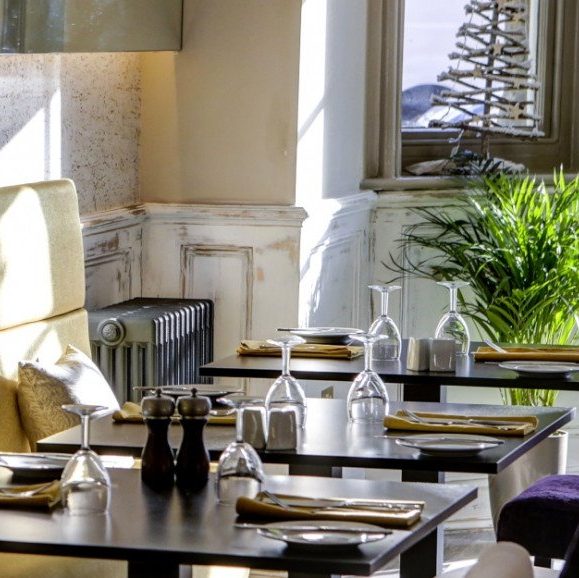 Sunlit restaurant dining area with neatly set tables, cushioned banquette seating, and a potted plant near the windows.