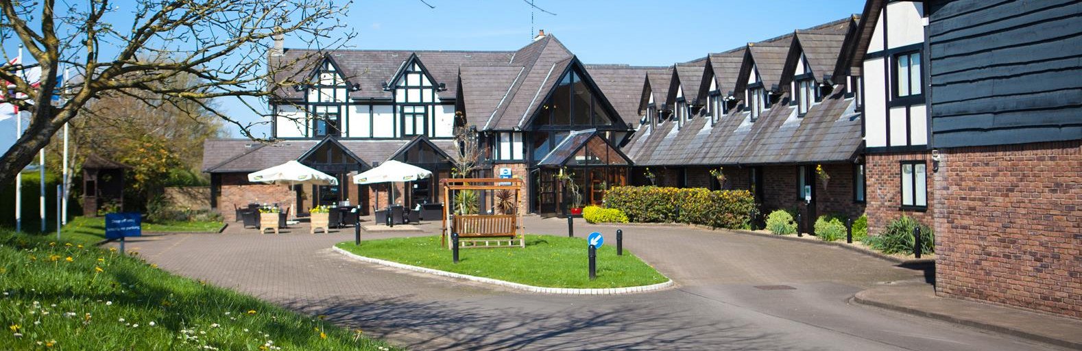 A large Tudor-style hotel building with a paved driveway, outdoor tables under white umbrellas, and a landscaped central island.
