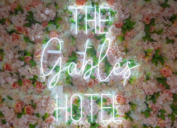 Neon text reading "The Gables Hotel" mounted on a wall covered with pink and white flowers and green leaves.