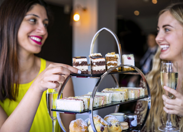 Two women smiling beside a tiered dessert stand with assorted cakes and pastries, one holding a glass of champagne.