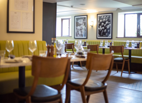 A softly lit restaurant interior with wooden chairs, set tables, green banquette seating, framed wall art, and windows along the back wall.