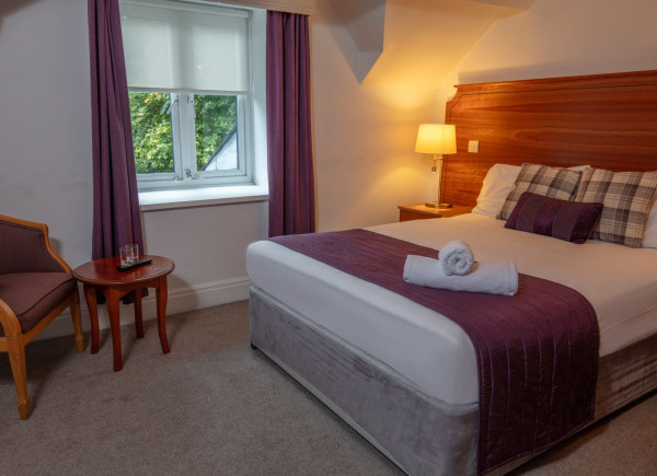 A neatly made bed with purple bedding and rolled towels in a bedroom with a window, lamp, chair, and small table.