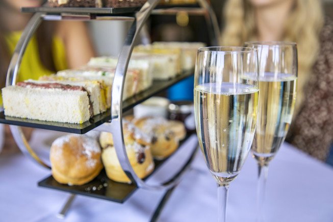Two champagne flutes in front of a tiered tray with sandwiches and pastries on a table.