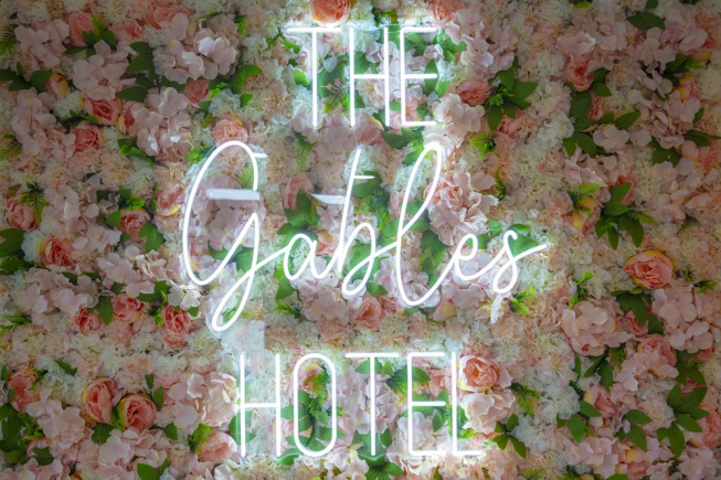 A neon sign reading "The Gables Hotel" glows over a dense wall of pink and white flowers.