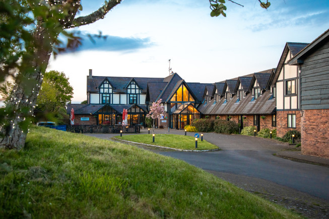 A lodge-style building with timber and brick фасades, lit windows, and a curved driveway in front of a grassy foreground at dusk.