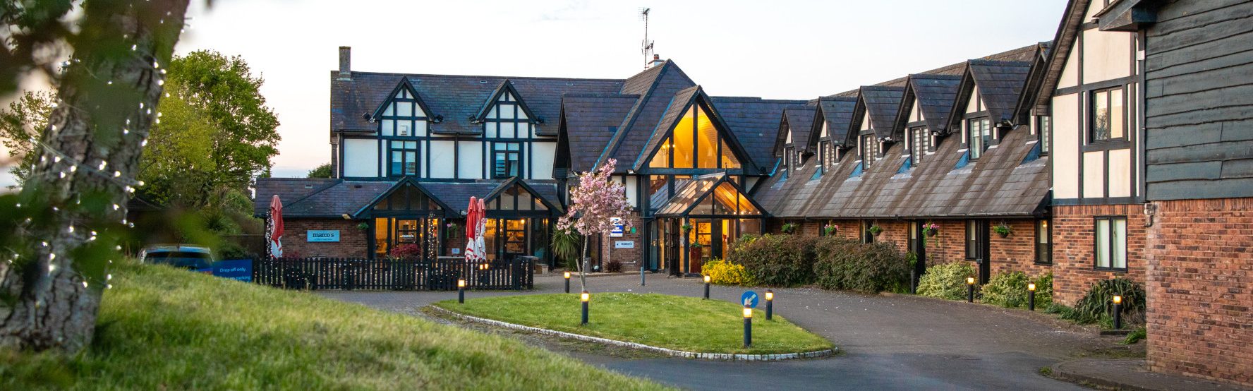 A large Tudor-style lodge or hotel building with lit windows, a central glass entrance, landscaped grounds, and a curved driveway at dusk.