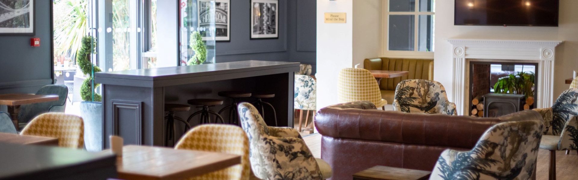 A stylish lounge with patterned chairs, small tables, a central dark bar or service island, and a fireplace on the right wall.