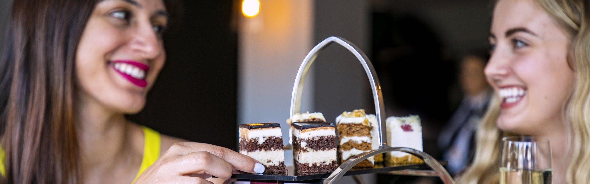 Two smiling women look at a tiered tray of cake slices in a restaurant or cafe.