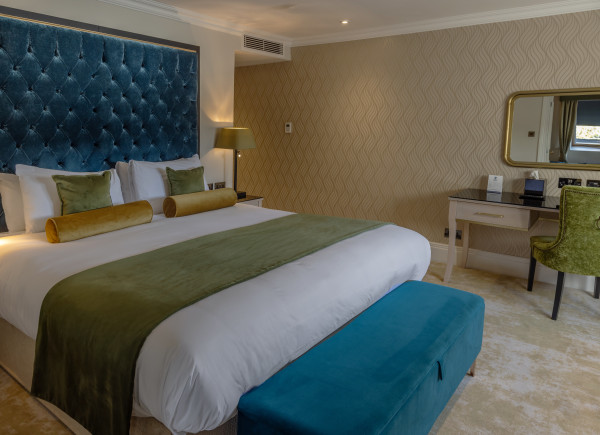 A hotel bedroom with a large bed, blue tufted headboard, green and white bedding, a teal bench, and a desk with a mirror and chair.