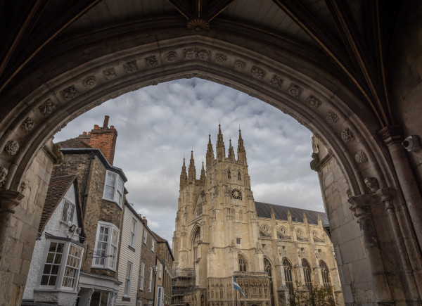 View of a large Gothic cathedral seen through a stone archway, with historic buildings on the left and a cloudy sky overhead.