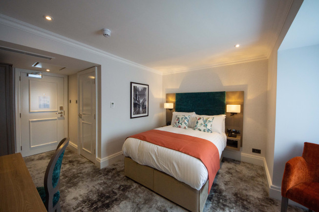 A modern hotel bedroom with a neatly made bed, bedside lamps, framed wall art, a desk chair, and a hallway door.