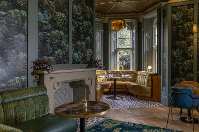 Interior of a decorated lounge with patterned wallpaper, a fireplace, booth seating by a window, and small tables.