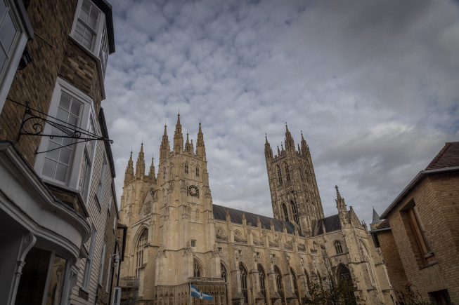 A large Gothic cathedral with tall spires rises between narrow buildings under a cloudy sky.