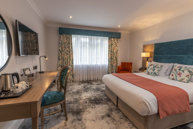 A modern hotel room with a large bed, desk and chair, wall-mounted TV, window with curtains, and a seating area.