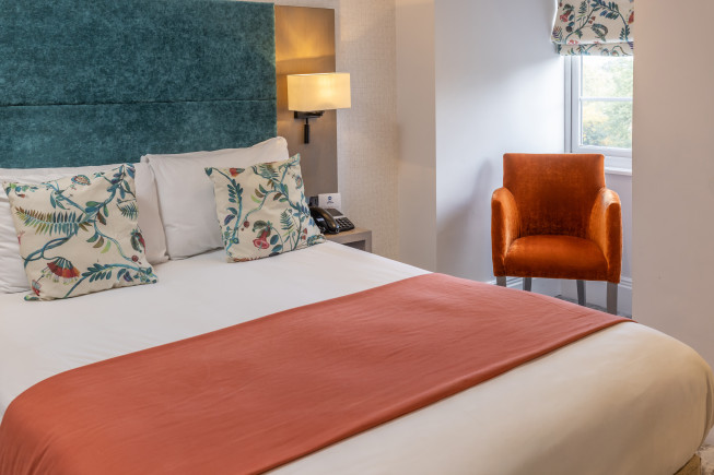 A hotel room with a neatly made bed, patterned pillows, an orange chair by the window, and a wall lamp beside the headboard.
